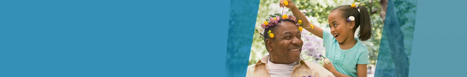 A smiling elderly man with a flower crown is playfully crowned by a cheerful little girl with flowers, against a green outdoor background.